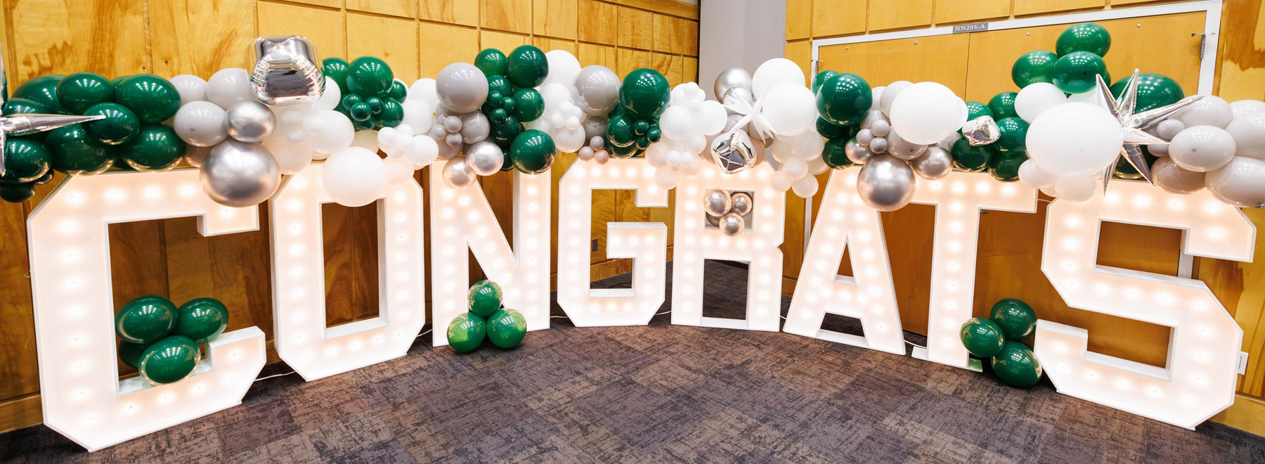 Large illuminated letters spelling ‘CONGRATS’ decorated with green, white, and silver balloons inside an event space.